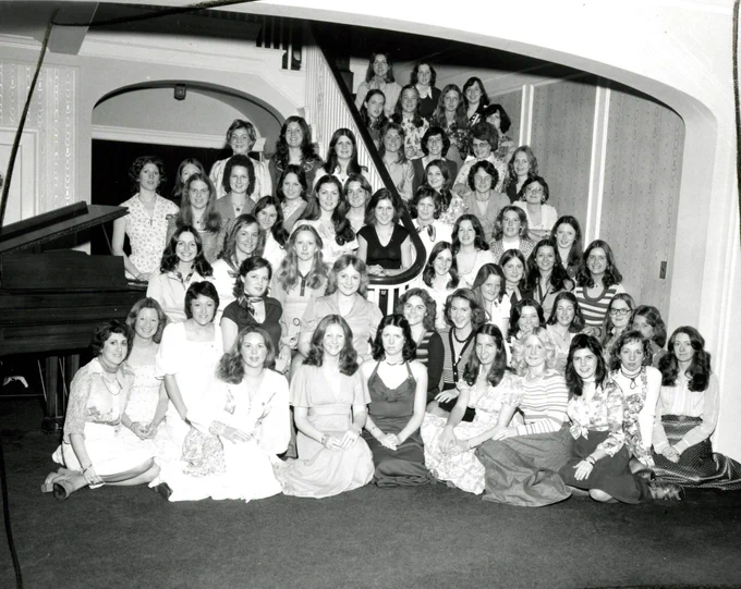 Black and white photograph from the WSU Libraries archives showing a large group of woman from Chi Omega sorority, posed on and around a staircase inside an ornate building. The group is arranged in multiple rows, with some seated on the floor and others standing on the steps. A grand piano is visible on the left side of the image, and the background features decorative architectural details, including arched openings and patterned walls. The individuals are dressed in mid-20th-century attire, such as dresses, blouses, and skirts.