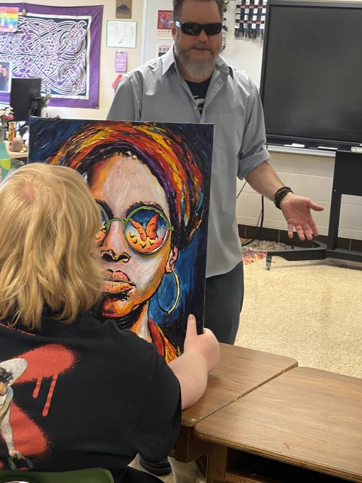 An ISBVI student sits at a table holding one of John Bramblitt’s vibrant, textured paintings, examining it closely. In front of the table, John Bramblitt is seen speaking to the class.