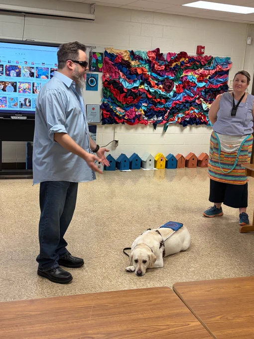 John Bramblitt is seen standing to the left of ISBVI art teacher Leslie Walsh as he addresses her art students. Bramblitt’s guide dog Zuke is laying on the ground between John and Leslie, with a colorful, textured art piece on the wall behind them.