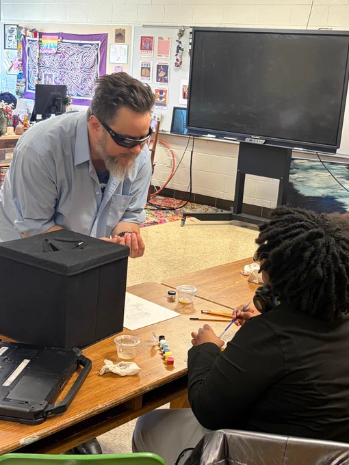 John Bramblitt stands near a table in Leslie Walsh’s art classroom as he speaks with a student who is drawing on foam paper. To the left of the student, there is a black box on the table, which holds an item that the student was drawing using only their sense of touch.