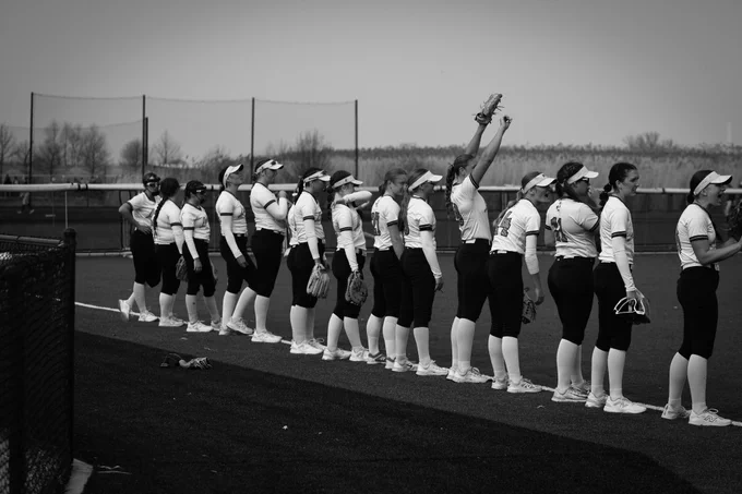 Fourteen Laker Softball players are lined up facing away from the camera, looking across the field. One player has her hands raised in excitement, and the other players are cheering. The photo is in black and white. 
