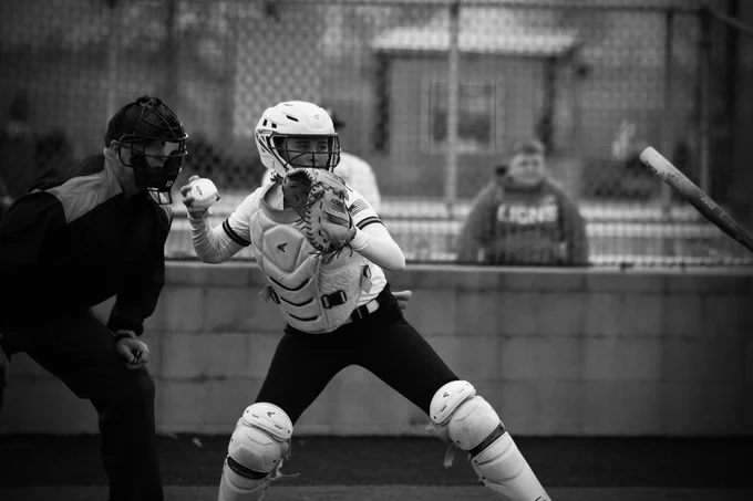 A Laker Softball player is wearing catching pads and a helmet. Behind her is the umpire while she prepares to throw the ball back to the pitcher. The photo is in black and white.