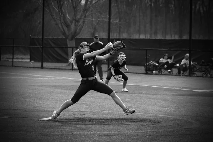 A Women's Laker Softball player is wearing a softball uniform while pitching a softball. Behind her is a player waiting to catch on first base. The photo is in black and white. 