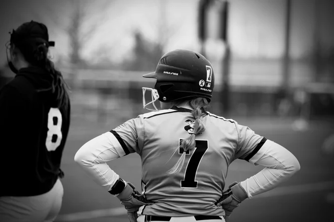 A Women's Laker Softball player has her back facing the camera while she looks off to the left. She is wearing a uniform and helmet, with the number 7 on the back. Her hair is in a braid and there is another player noticeable wearing the number 8. The photo is in black and white. 

