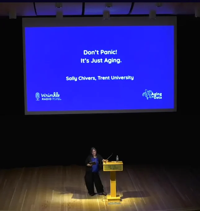 Photo of Dr. Sally Chivers (Trent University) delivering the Centre on Aging and Health Distinguished Public Lecture, "Don't Panic! It's Just Aging." on a stage at the University of Regina's Education Auditorium. A screen with the lecture topic and logos for "Wrinkle Radio" and "Aging in Data" are displayed behind the speaker.
