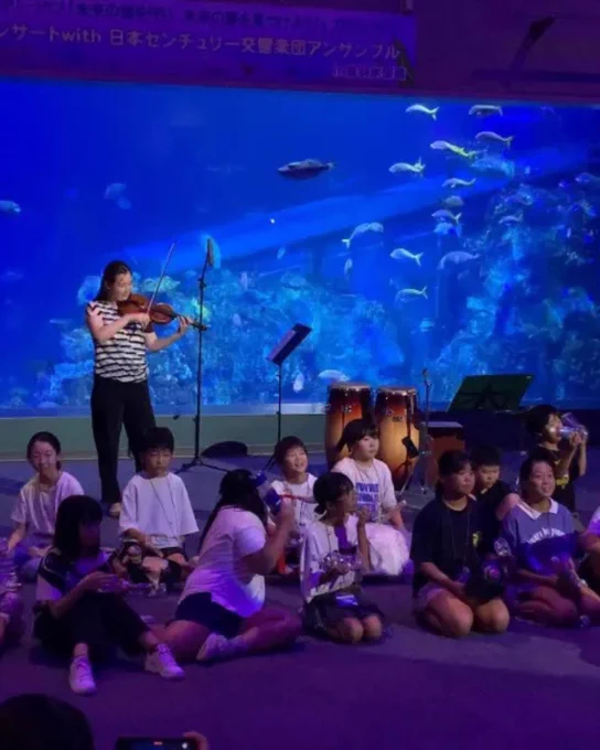 A group of people seated on the floor in front of a large aquarium display filled with fish. One person is standing and playing a violin. Some percussion instruments are visible in the background.