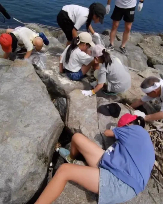 A group of individuals is gathered on large rocks near a body of water. They seem to be searching through the rocks, possibly looking for items or conducting a study. Some people are wearing hats, and a few are crouched down, focused intently on their tasks. The scene suggests an outdoor activity or exploration.