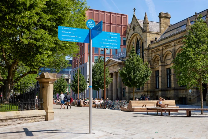 A city plaza with a blue directional sign in the foreground, indicating different locations. University buildings are in the background, surrounded by trees and benches. The area is paved and well-lit, with bicycles parked near the buildings.