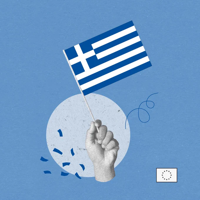 A raised hand holds a small Greek flag against a blue background, with scattered confetti and a tiny European Union flag in the corner suggesting a celebratory European context.