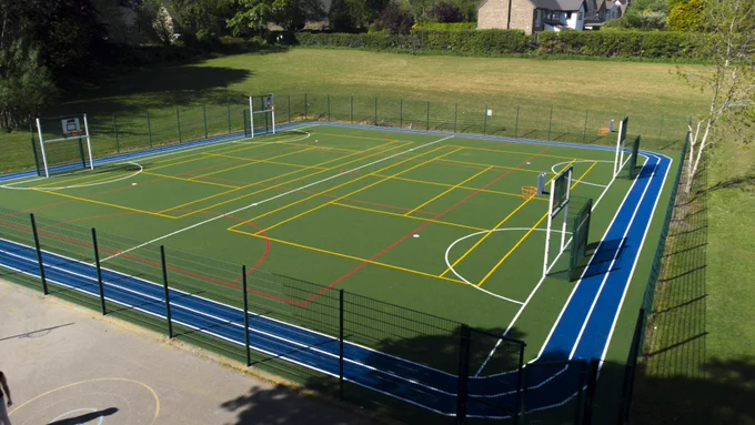 Outdoor multi-sport court with basketball hoops and various colored lines, surrounded by a blue running track and black fencing.