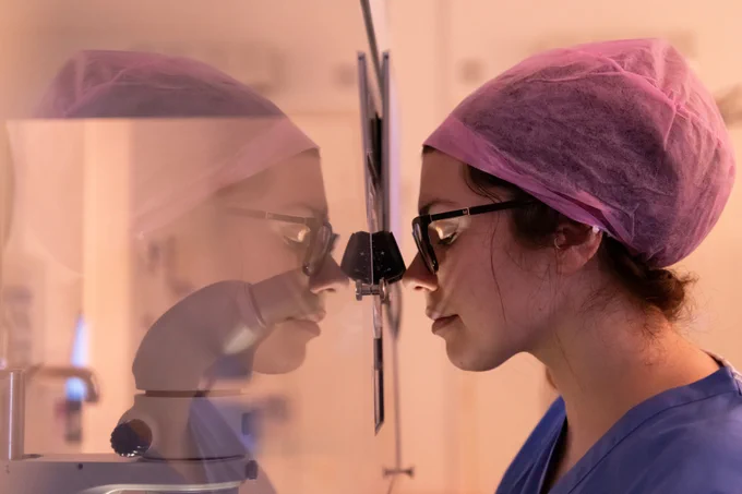 Close up photo of a female researcher looking through a microscope in a clinical setting. She is wearing blue hospital scrubs and a pink hair net.