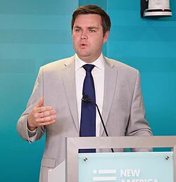 "JD Vance, bearded man in a dark suit and red tie, standing at a podium with a serious expression, American flag in the background during a political speech.