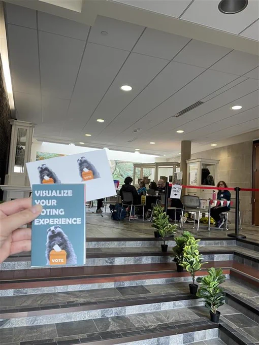 Hand holding voting experience cards with a badger voting image inside UW School of Human Ecology in-person absentee voting site with seated voters and staff.