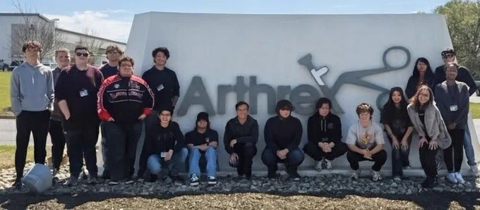 A group of 18 people is posing for a photo in front of a large Arthrex sign, featuring the company logo and name in large, gray letters.