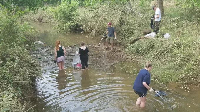 Five people pick up and bag trash in a wide creek and along the banks