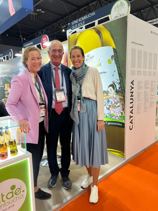 Tres personas sonrientes posan en un stand de feria junto a un expositor de kombucha y aceite.