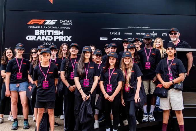 A group of students attending the Formula 1 Qatar Airways British Grand Prix 2025 pose together in front of an event display, wearing matching caps and passes.