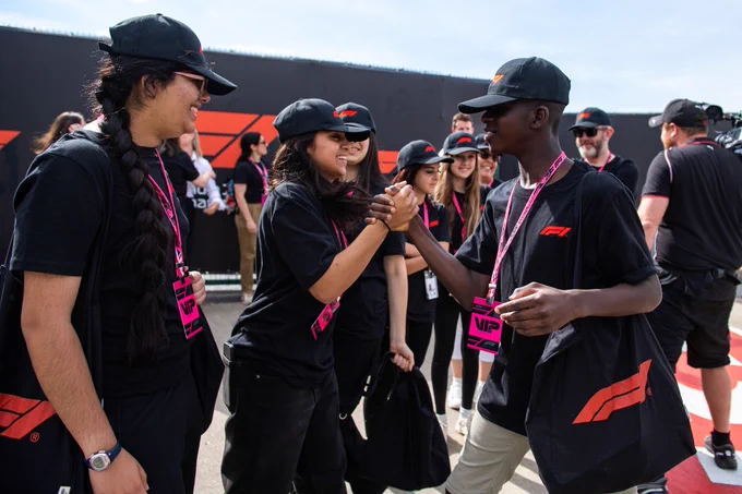 Students at the Formula 1 Qatar Airways British Grand Prix 2025 greet each other in an outdoor trackside setting, wearing Formula 1 caps and lanyards.