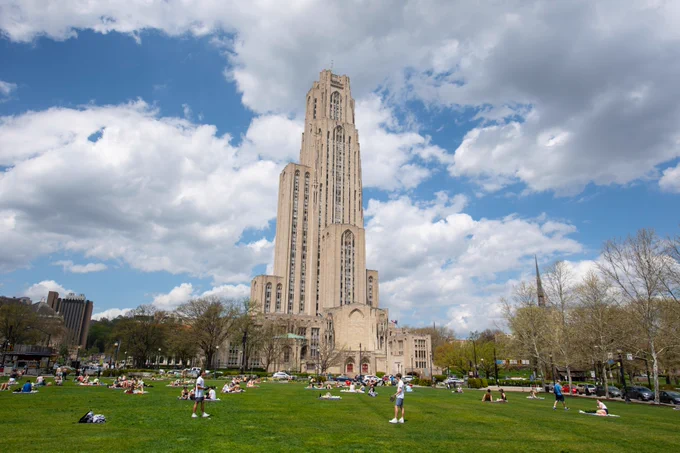 The image shows the Cathedral of Learning in Pittsburgh, framed by a blue sky with scattered clouds. People are relaxing on a large grassy area in the foreground.