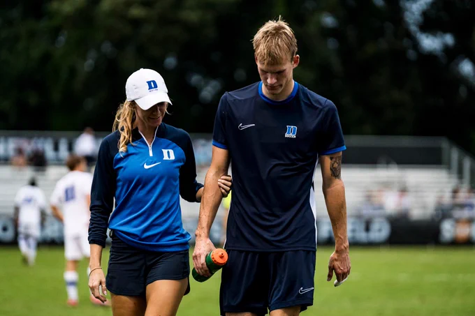 A female trainer in a blue and black outfit assists a male soccer player wearing a dark uniform. The player, holding a water bottle, appears to be walking off the field with her support. In the background, other players are seen on the field during a soccer match. Trees are visible in the distance.