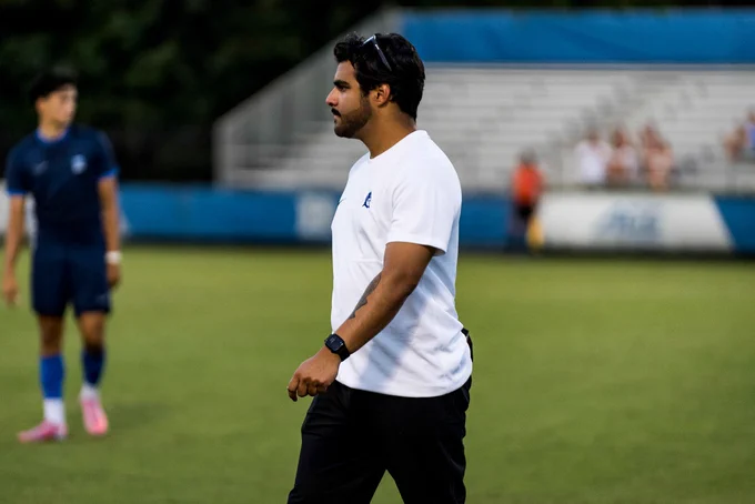 A coach in a white shirt and black pants walks along the field, observing a soccer practice. In the background, players can be seen on the field and some spectators in the stands.