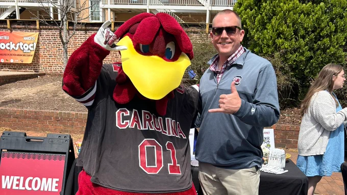 A person is posing with the University of South Carolina's mascot, who is wearing a jersey with "CAROLINA" and the number "01". They're standing outdoors with displays behind them, and the mascot is giving a spurs-up.