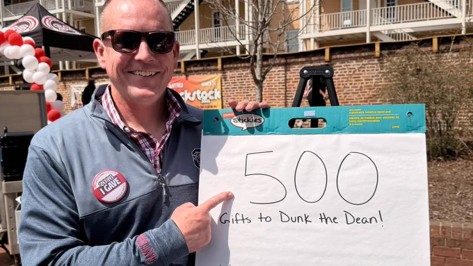 Dean Hodges standing outdoors next to a flip chart with a handwritten note that says "500 Gifts to Dunk the Dean!" with a list of numbers below. They're wearing a University of South Carolina button and sunglasses, and they are smiling while pointing at the chart. There are balloons and a building in the background.