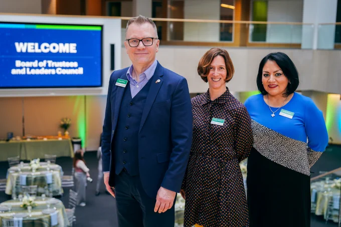 Joseph Hafner, Helen Thomas, and Lorely Ambriz, members of the OCLC Leaders Council, stand in front of a screen displaying \"Welcome OCLC Board of Trustees and Leaders Council.\" Tables with floral arrangements are in the background.