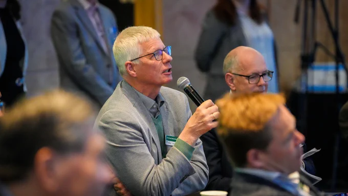 Reinhard Altenh\u00F6ner, Deputy General Director of Staatsbibliothek Berlin and member of the OCLC Leaders Council, speaks into a microphone. Reinhard is wearing glasses and a gray suit jacket. Other attendees are visible in the blurred background.