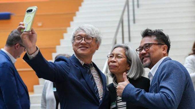 Three people smile as they pose for a selfie in front of a staircase during the gathering of the OCLC Leaders Council. The person on the left holds up a cell phone, and the person on the right gives a thumbs-up.