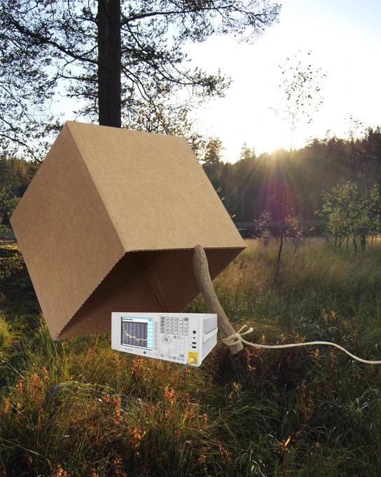 A large cardboard box propped up with a stick like a classic trap sits in a grassy field at sunset. Under the box is an oscilloscope.