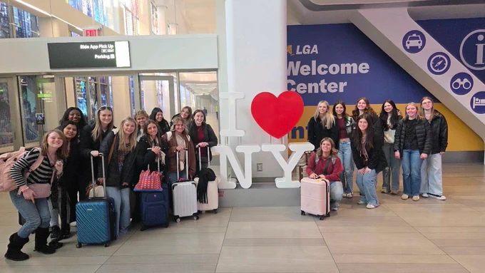 A group of young women travelers pose with their suitcases around a large "I ❤️ NY" sign at the LaGuardia Airport (LGA) Welcome Center. The group is split on either side of the iconic white letters and red heart, with modern airport signage and glass doors visible in the background.
