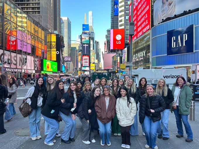 A large group of young women stands together in the heart of Times Square, New York City. They are positioned in the foreground of a wide pedestrian area, surrounded by the iconic bright digital billboards for brands like GAP, LG, and Broadway's The Lion King. The group is dressed in casual winter layers, and the bustling city streets and skyscrapers extend into the background under a clear sky.