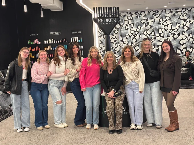A group of nine women pose together at the Redken Exchange salon academy. They are standing in front of a large, decorative black hair pick and oversized silver scissors. The background features shelves of hair products and a black-and-white patterned wall. The group is dressed in stylish, casual outfits including sweaters, jeans, and boots.