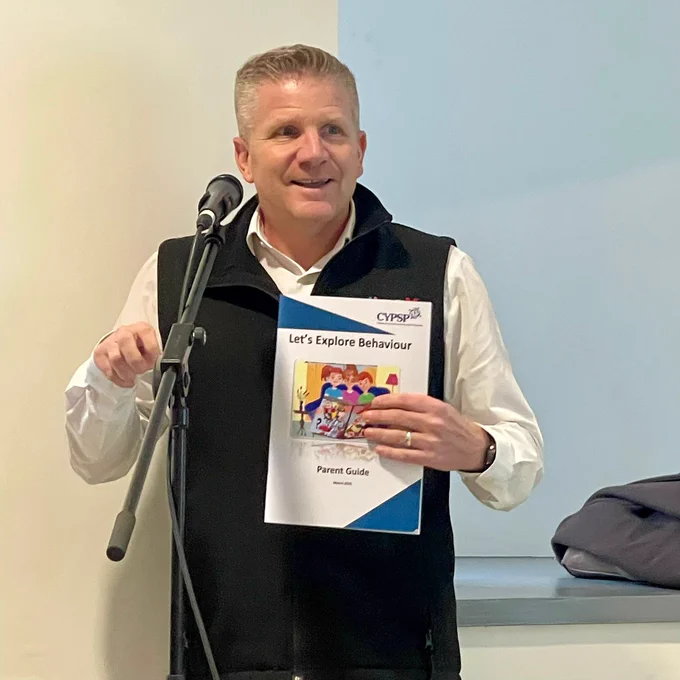 Person standing at a microphone holding a booklet titled ‘Let’s Explore Behaviour – Parent Guide