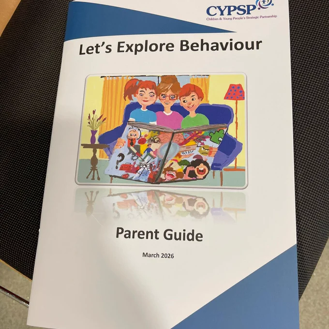 A close-up of a booklet titled ‘Let’s Explore Behaviour – Parent Guide,’ featuring an illustrated cover showing three individuals sitting on a sofa reading a large picture book