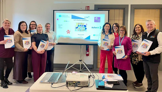 A group of people standing in front of a presentation screen, each holding the Lets Explore Behaviour Parent Guide
