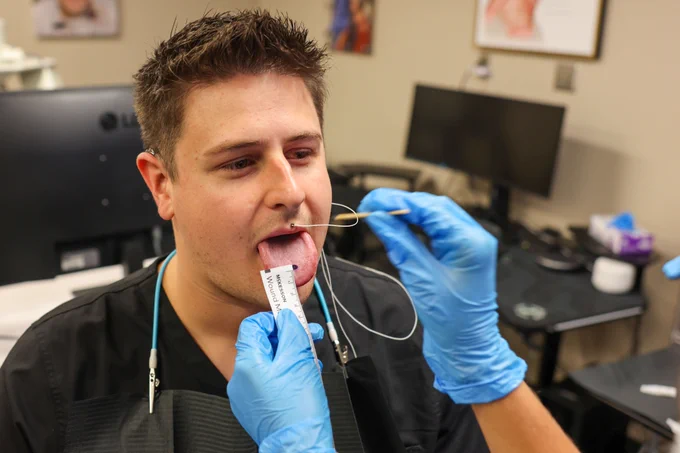 A person with short hair sits in a medical office, sticking out their tongue. Clad in blue gloves, a medical professional uses a tool to apply a small white strip labeled "Pfizer" to the tongue. The room features a computer, medical equipment, and framed pictures on the wall.
