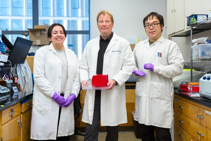 Three researchers in white lab coats holding in a laboratory, standing in front of blue-windowed workspace holding a piece of equipment.