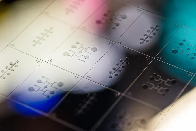 An array of circuit-like patterns on a metal wafer, illuminated in blue, pink, and yellow light.