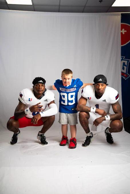 Maddox in a blue West Georgia football jersey posing with two UWG football players in front of a white backdrop