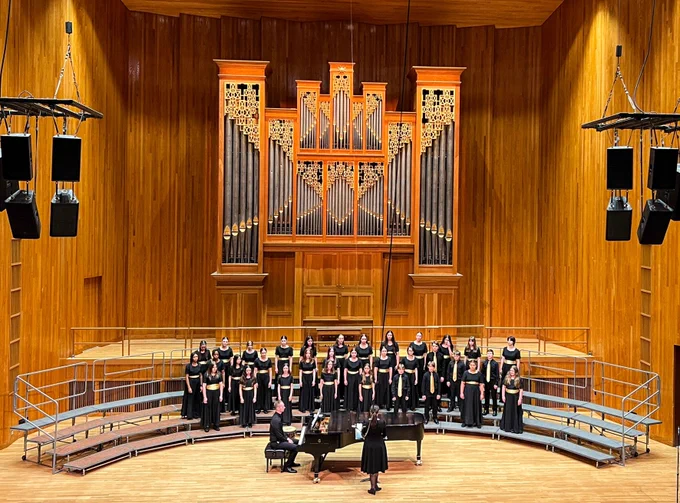 A choir performing on a large wooden stage, accompanied by a pianist. Above them is an impressive pipe organ. The choir members stand in semi-formal attire, arranged on tiered platforms. Two grand pianos are on the stage.
