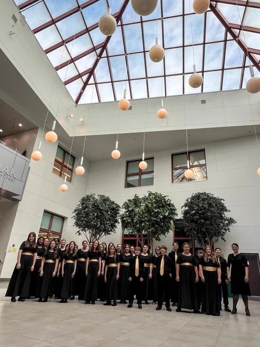 A group of people dressed formally stand together in an atrium with a high, glass-paned ceiling and spherical hanging lights. Two large potted trees are behind the group, and windows line the white walls.