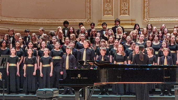 A large choir performs at Carnegie Hall. The group stands on stage behind a grand piano, all wearing coordinated black attire with gold accents. The ornate backdrop features intricate detailing.