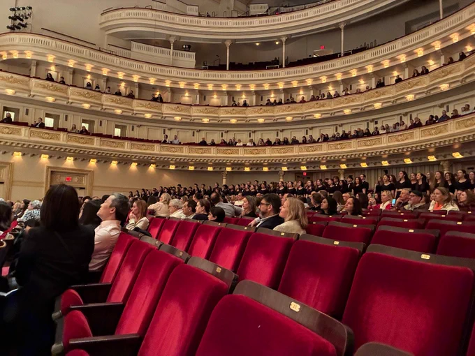 A large audience seated inside Carnegie Hall, view facing the stage, showing multiple tiers of balcony seating and ornate architectural details.