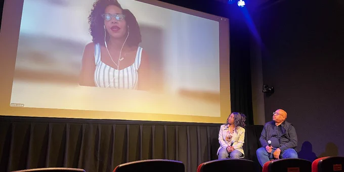 Two people seated on stage looking up at a large screen displaying a woman during question and answer segment of documentary screening