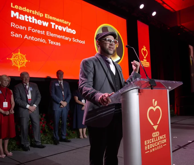 A Texas A&M University-Corpus Christi alumnus speaks at a podium on a stage with a large screen behind displaying "Leadership Elementary, Matthew Trevino, Roan Forest Elementary School, San Antonio, Texas." Several people are standing in the background. The event branding shows "H-E-B Excellence in Education Awards.