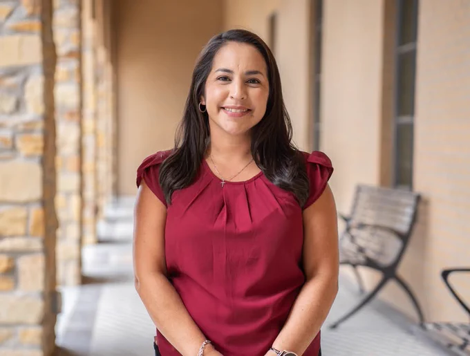 A smiling administrative leader and Texas A&M University-Corpus Christi alumna stands in a corridor with stone walls and a bench. They are wearing a maroon blouse.
