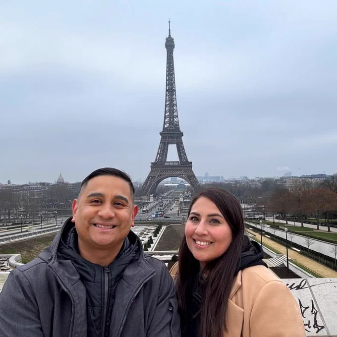 A Texas A&M University-Corpus Christi alumna and her husband pose for a selfie photo smiling in front of the Eiffel Tower in Paris. The sky is overcast, and there are pathways and trees in the background.