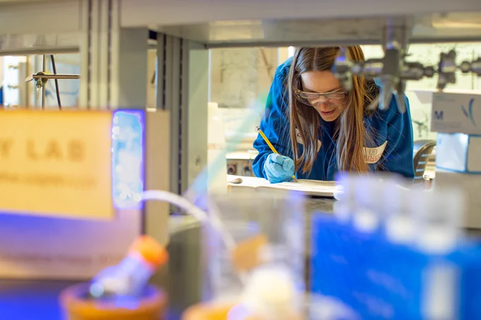 A scientist wearing a blue lab coat and gloves records notes in Christina Cooley's lab.